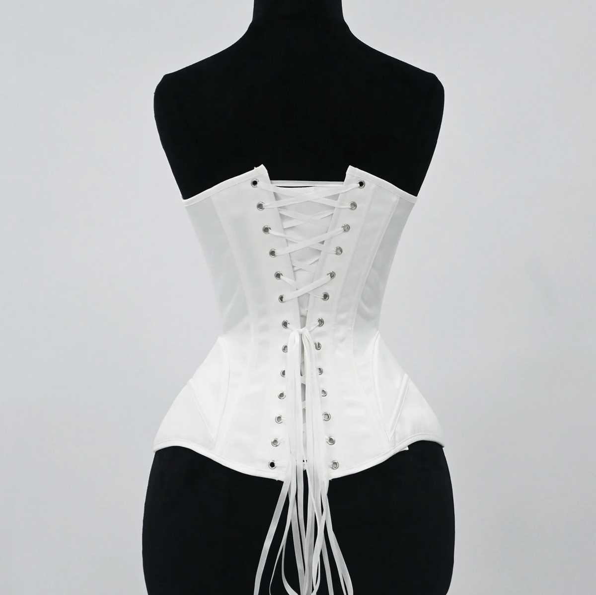 White satin bridal corset featuring a structured lace-up back with silver grommets and long ribbon ties on a mannequin.