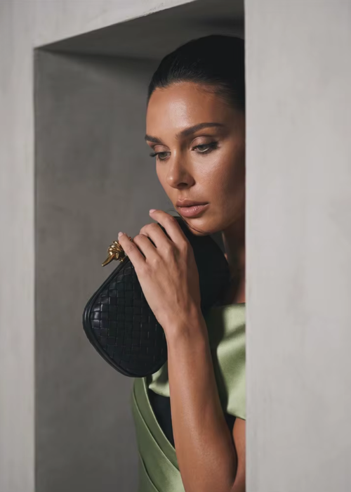 Woman wearing a structured sage green satin twill gown with a black bodice, holding a woven black clutch with gold detail.