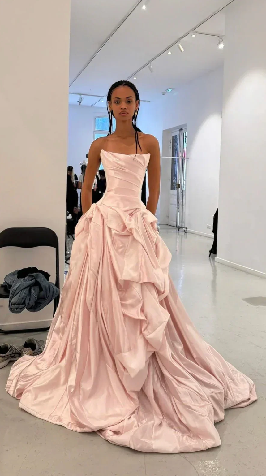 Model in a blush pink satin off-shoulder A-line gown with a pleated bodice and voluminous, draped floor-length skirt.