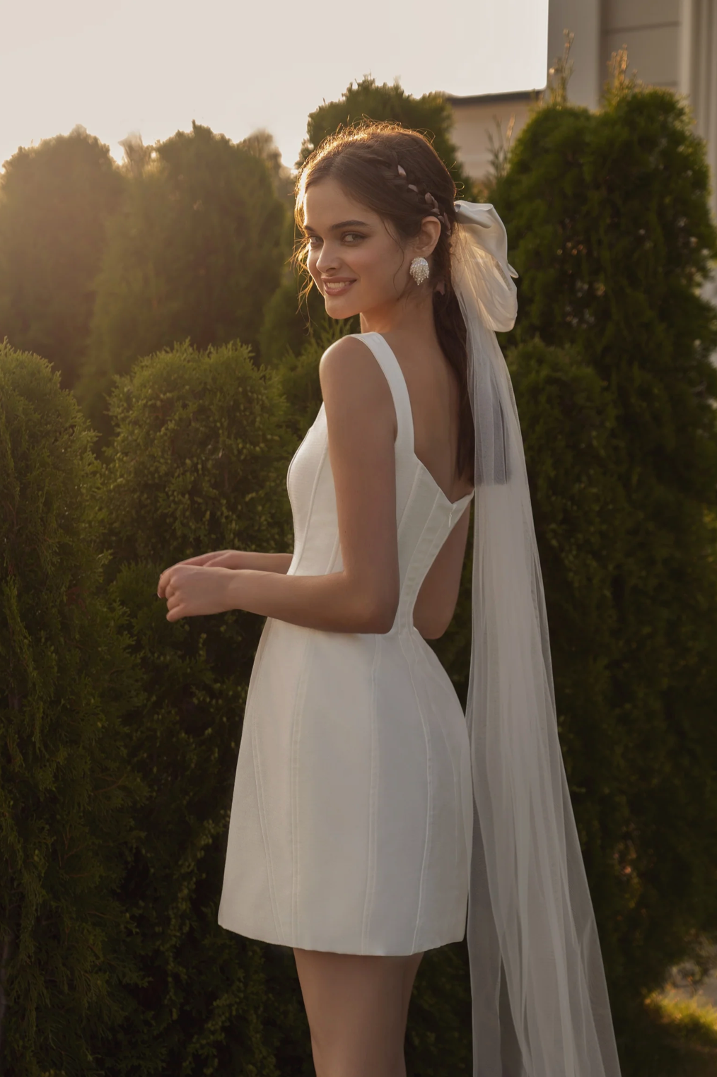 White structured bridal mini dress with a square neckline, corset bodice, and thin straps, worn with a long veil.