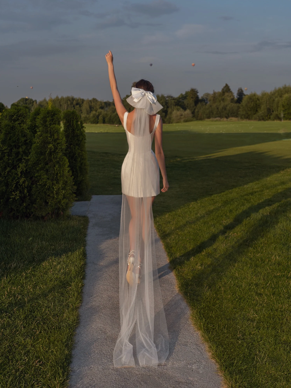 Hilda white bridal mini dress with a structured corset bodice, square neckline, and long sheer veil on a grassy path.