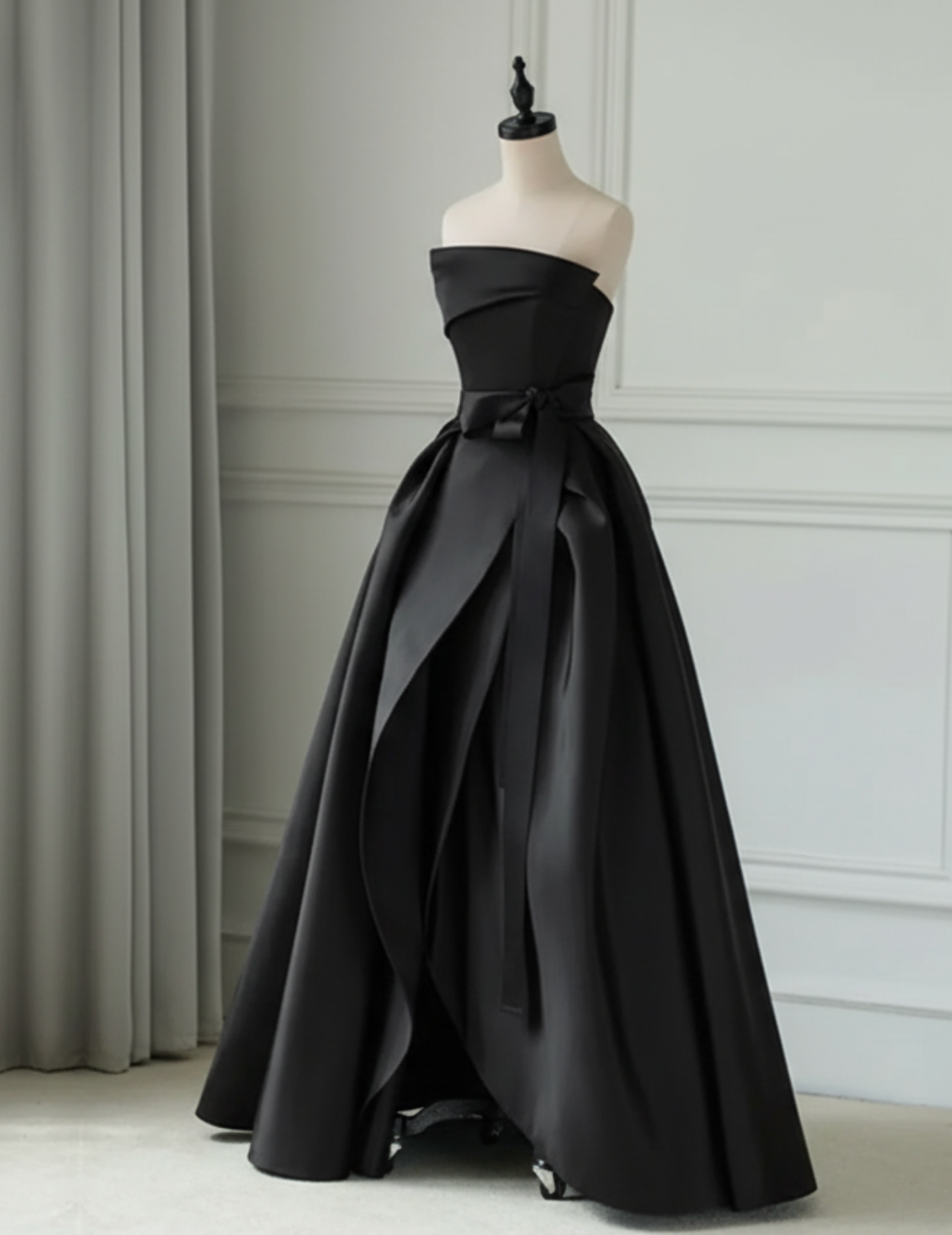 Black strapless satin twill gown with a pleated A-line silhouette, ruched bodice, and elegant waist sash on a mannequin.
